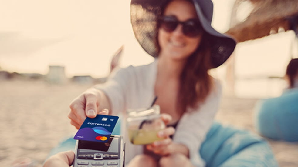 Woman sat on sandy beach, wearing floppy sun hat and sunglasses, she holds a drink in one hand and extends her other arm and hand out, as she holds a payment card to a single card payment machine. 